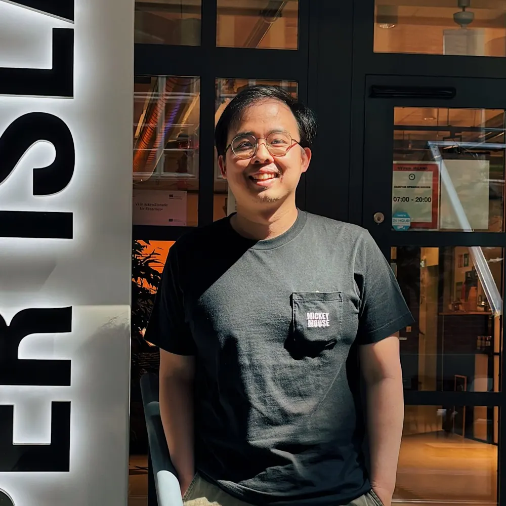 Thomas Tjaja, tersenyum, berdiri di depan tanda Hyper Island